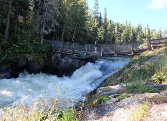 Una persona de pie en el puente colgante del Parque Provincial de las cataratas Wekusko, viendo pasar el agua.