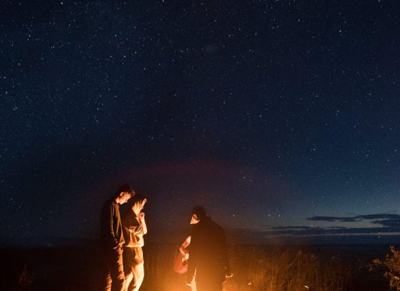 Vier Personen stehen um ein Lagerfeuer mit Blick auf den nächtlichen Wald.