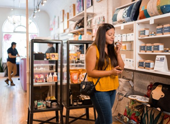 Una persona con camisa amarilla huele una pequeña vela frente a hileras de productos locales en Tara Davis Studio Boutique.