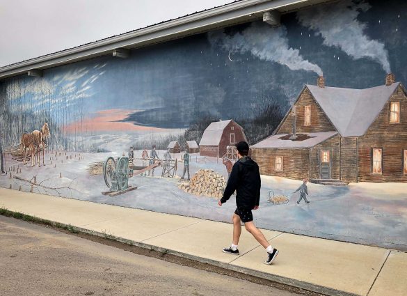A person walks by a mural showing a winter scene of a house, barn and people working with logs.