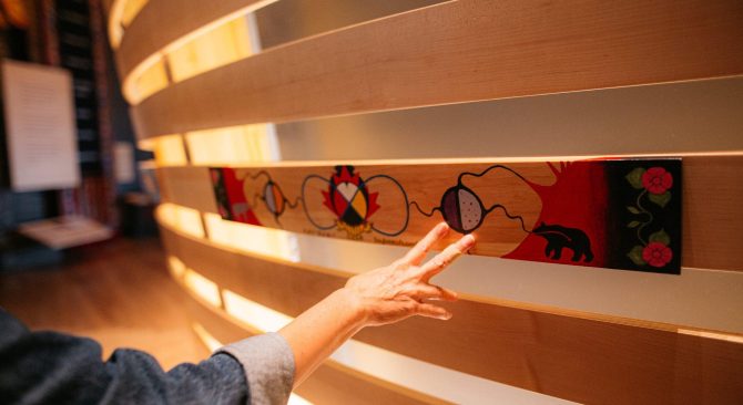 Una mano toca una larga pieza de madera pintada con coloridos diseños, entre ellos una rueda medicinal y flores de estilo mestizo.