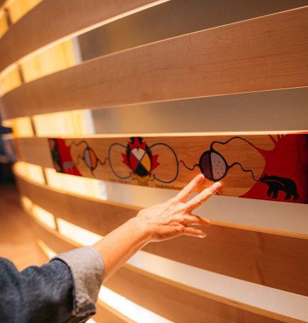 Une main touche une longue pièce de bois peinte de motifs colorés, dont une roue médicinale et des fleurs de style métis.