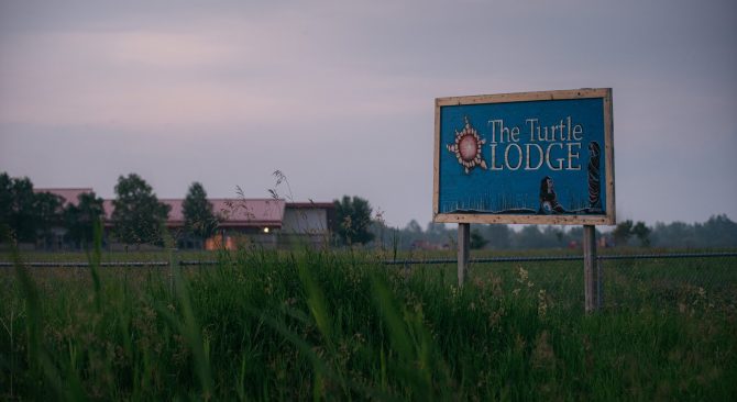 Un cartel azul que dice "The Turtle Lodge" en un campo verde al atardecer, un edificio a lo lejos en el fondo.