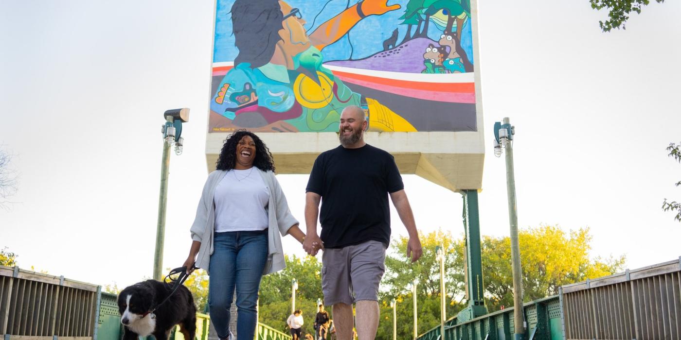 A couple holding hands and walking a dog passes under the colourful mural on the footbridge at The Forks.