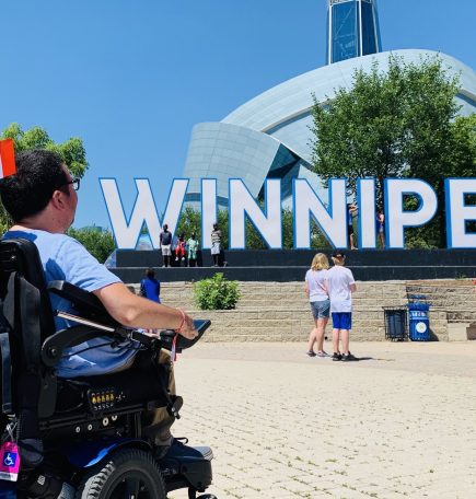Person in a wheelchair looking at the CMHR behind the Winnipeg sign at The Forks.