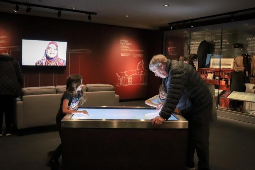 Grandpa & grandchildren captivated by interactive digital displays at the Winnipeg Museum, sharing moments of exploration.