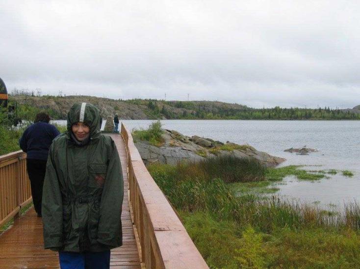 Taong naglalakad pabalik sa basang kahoy na Flin Flon boardwalk na nakasuot ng raincoat at nakahood pagkatapos ng mother nature rain shower.
