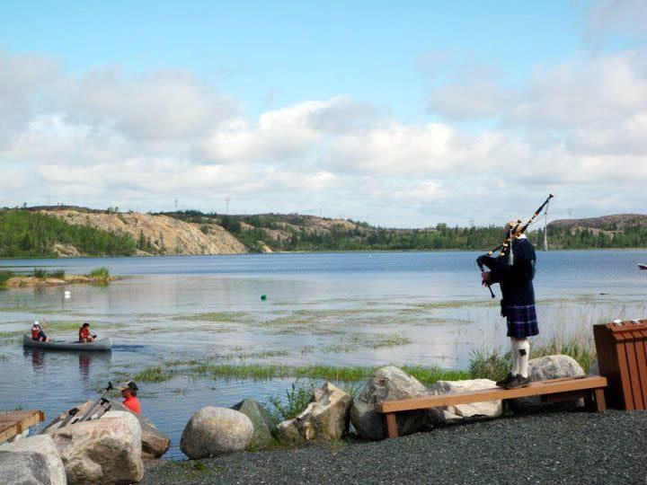 Tinatanaw ng bagpiper sa bench ang tubig habang dumadausdos ang mga canoeist sa gilid ng baybayin.