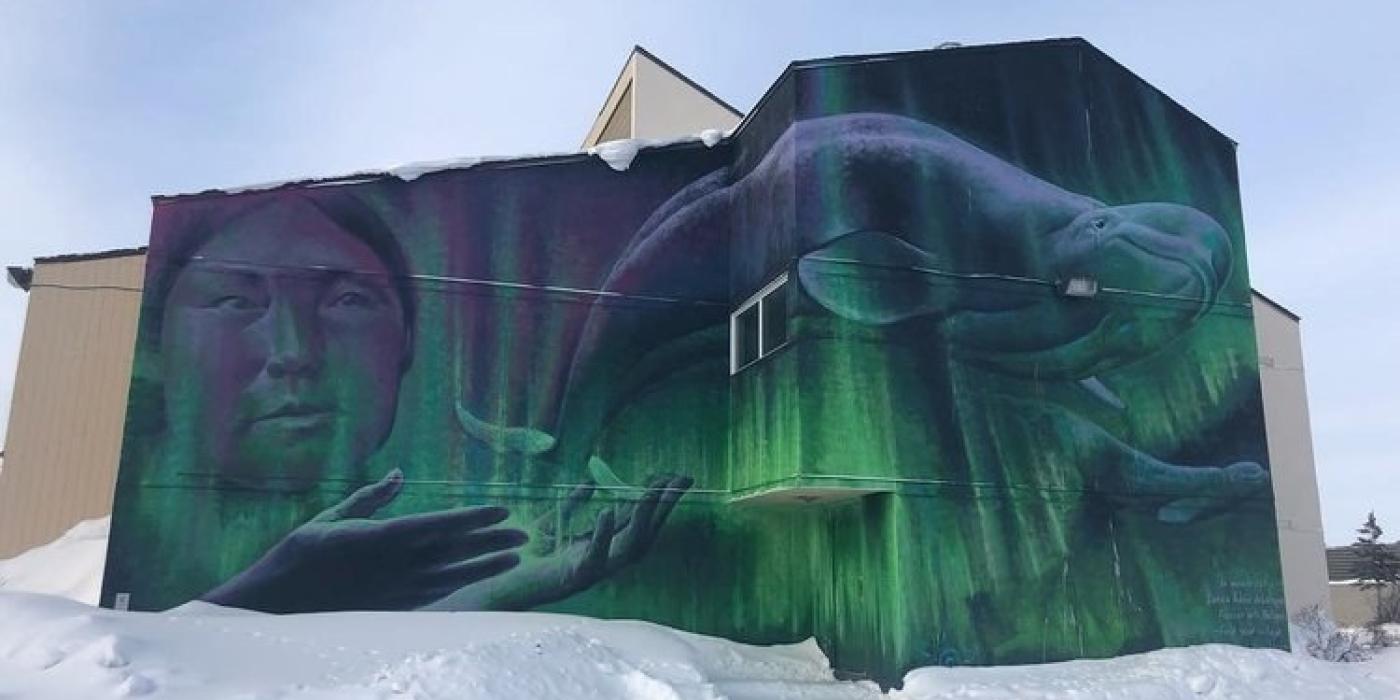 Mural of northern lights overlaid with a persons face and a beluga whale, painted on the side of a building in Churchill.