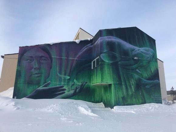 Wandgemälde von Nordlichtern, überlagert mit dem Gesicht einer Person und einem Belugawal, gemalt an der Seite eines Gebäudes in Churchill.