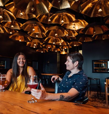 Two people drink beer from glasses inside of Nonsuch Brewing under a ceiling of gold umbrellas.