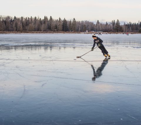 Person beim Schlittschuhlaufen mit einem Hockeyschläger auf einem glasartig gefrorenen See im Riding Mountain National Park.