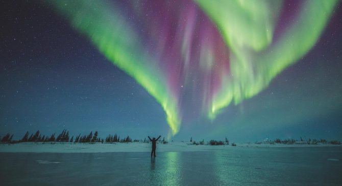 Person on a frozen lake with arms stretched to the sky under the purple and green northern lights.