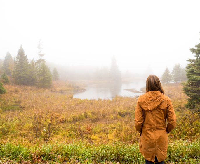 A person standing at the edge of a marsh, looking out over the fog covered water.
