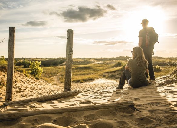 Una persona sentada y otra de pie junto a los postes de una valla de madera, mirando hacia las dunas en un día soleado.