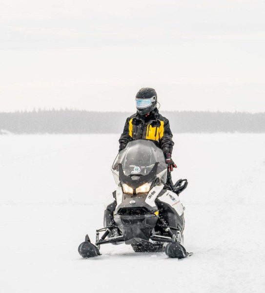 Una persona de pie sobre una moto de nieve cruzando un espacio nevado abierto.
