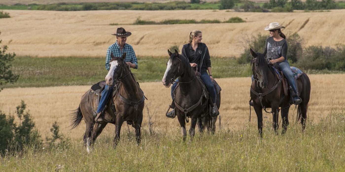 Tres personas a caballo en lo alto de una colina con campos de trigo al fondo.