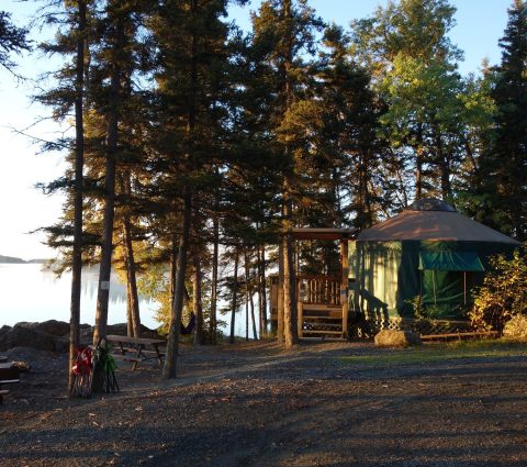 Una yurta junto a un lago, rodeada de árboles y con una mesa de picnic en primer plano.