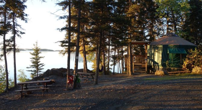 Una yurta junto a un lago, rodeada de árboles y con una mesa de picnic en primer plano.