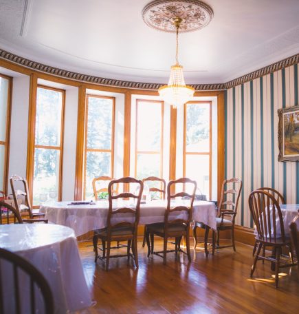 Un luminoso comedor con muchas ventanas y suelos de madera, que alberga cuatro mesas cubiertas con manteles y sillas blancas.