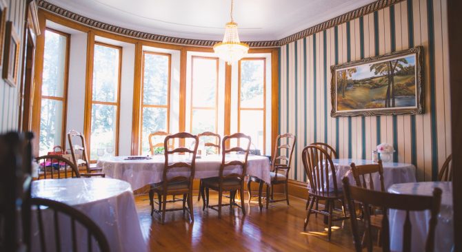 Un luminoso comedor con muchas ventanas y suelos de madera, que alberga cuatro mesas cubiertas con manteles y sillas blancas.