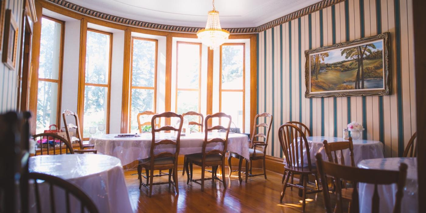 Un luminoso comedor con muchas ventanas y suelos de madera, que alberga cuatro mesas cubiertas con manteles y sillas blancas.