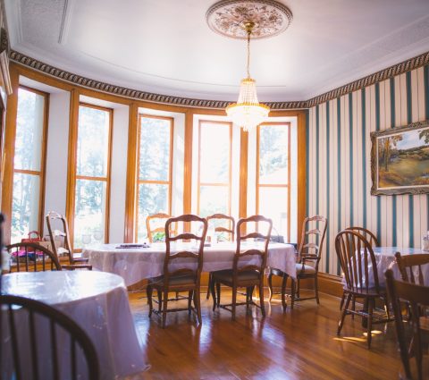 Un luminoso comedor con muchas ventanas y suelos de madera, que alberga cuatro mesas cubiertas con manteles y sillas blancas.