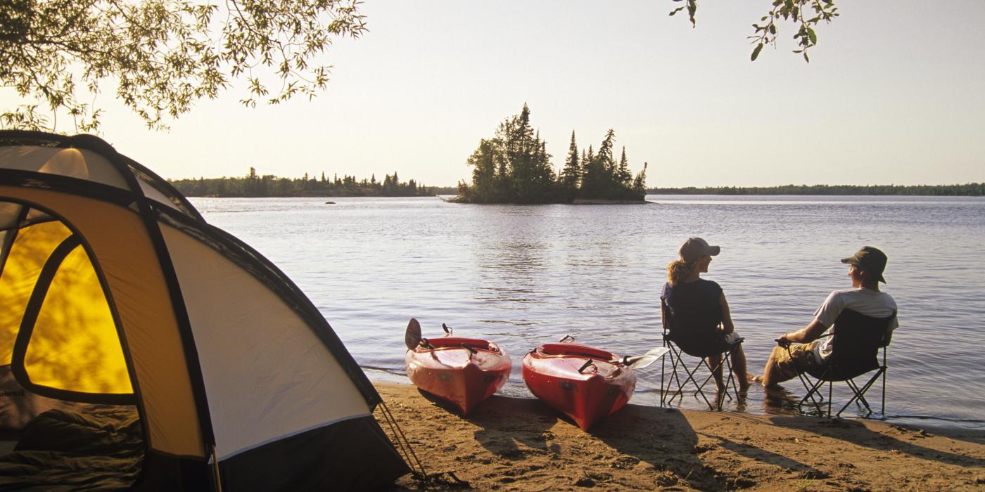 Two people in camping chairs sit next to their kayaks in front of their tent on the shore of a lake in the Whiteshell.