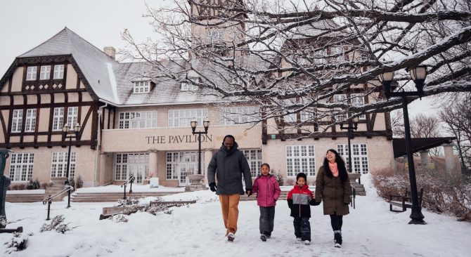 Une famille composée de deux enfants et de leurs deux parents se promène devant le pavillon Assiniboine Park à Winnipeg en plein hiver.