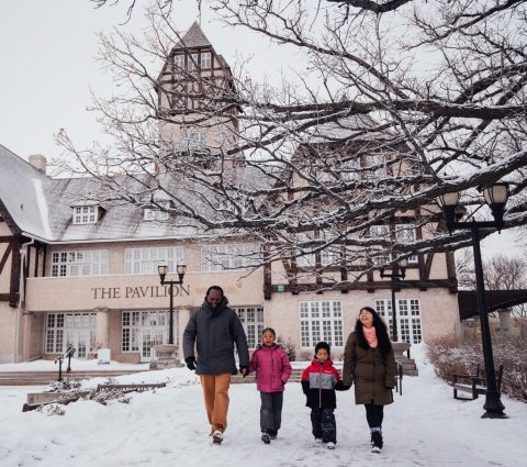 Une famille composée de deux enfants et de leurs deux parents se promène devant le pavillon Assiniboine Park à Winnipeg en plein hiver.
