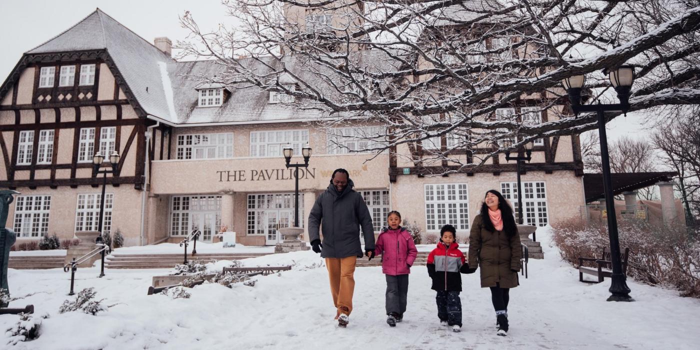 A family of two kids and two parents walks outside in winter in front of the Assiniboine Park Pavilion building in Winnipeg.