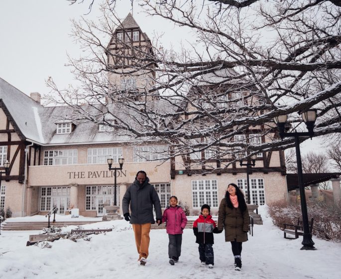 Une famille composée de deux enfants et de leurs deux parents se promène devant le pavillon Assiniboine Park à Winnipeg en plein hiver.