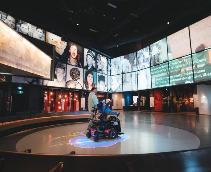 An exhibition hall with large screens showing diverse faces and freedom-themed text; a visitor uses a mobility scooter.