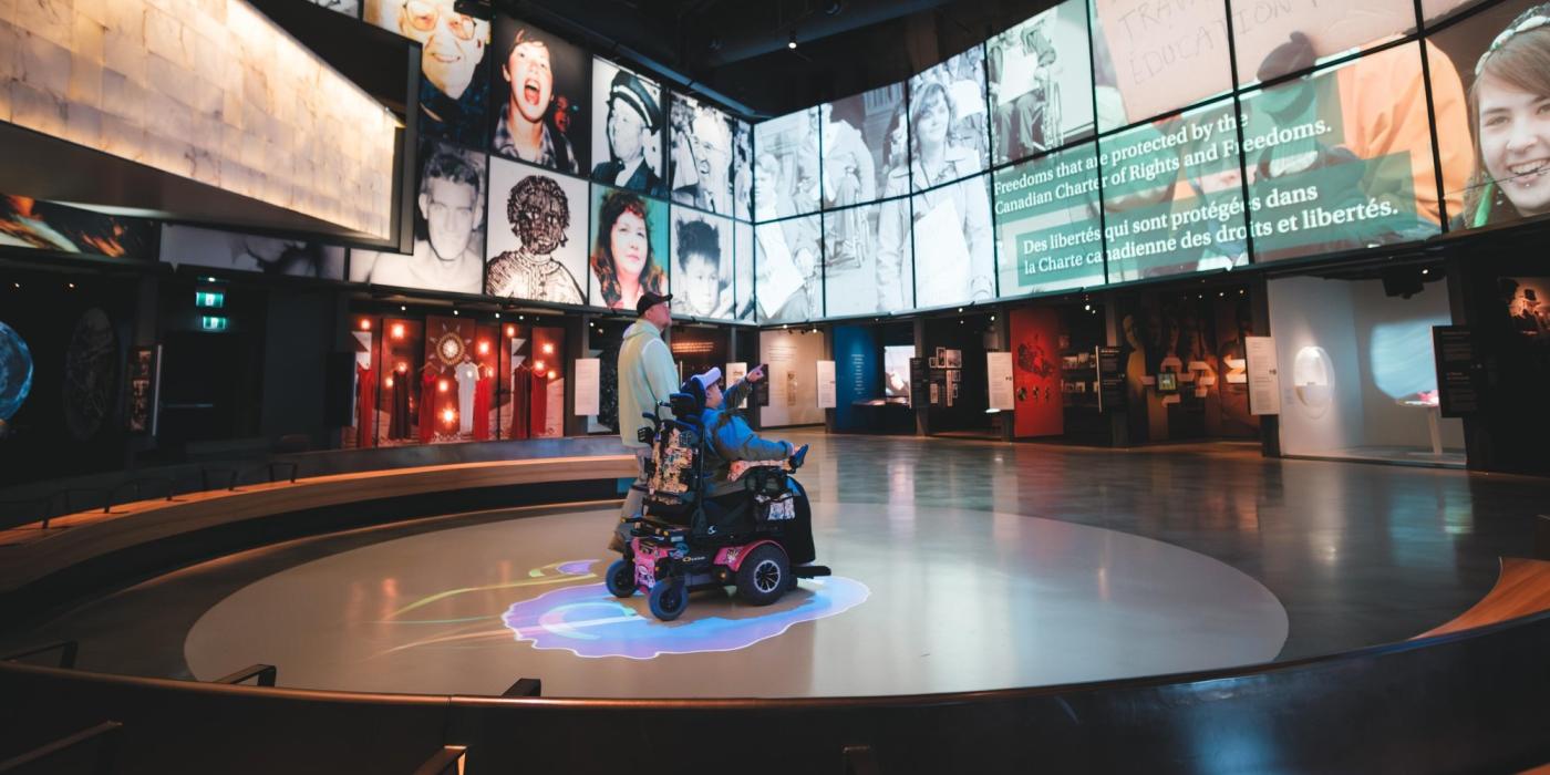 An exhibition hall with large screens showing diverse faces and freedom-themed text; a visitor uses a mobility scooter.