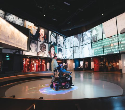 Une salle d'exposition équipée de grands écrans sur lesquels défilent des visages variés et des textes sur le thème de la liberté ; un visiteur se déplace à bord d'un scooter électrique.
