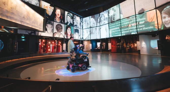 Une salle d'exposition équipée de grands écrans sur lesquels défilent des visages variés et des textes sur le thème de la liberté ; un visiteur se déplace à bord d'un scooter électrique.