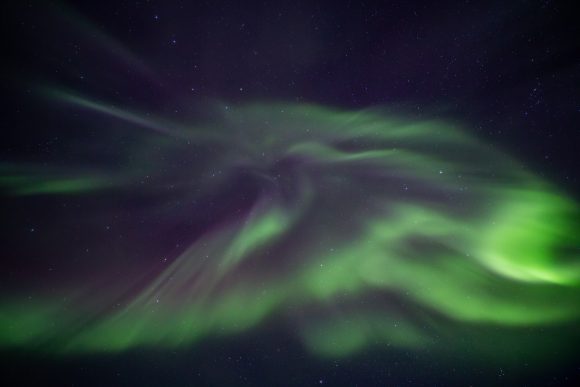A dense, swirling display of green and violet aurora borealis fills the night sky, with faint stars visible.