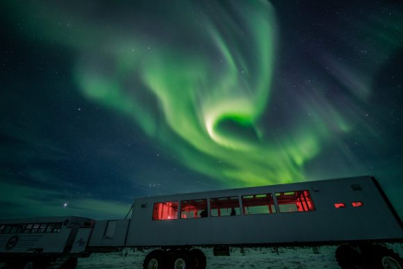 Green aurora borealis swirls above a parked tundra buggy vehicle with red-lit interior windows, against a starry night sky.