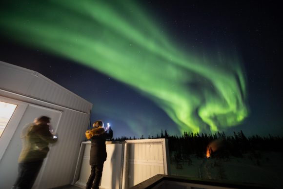 People with phone cameras stand near a cabin as sweeping green aurora borealis fills the sky above snowy trees.