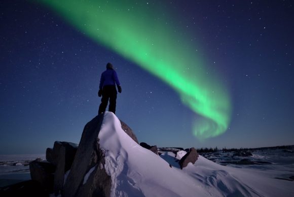 A person stands atop a snowy rocky point as a single green aurora borealis arc streaks across the starry sky.