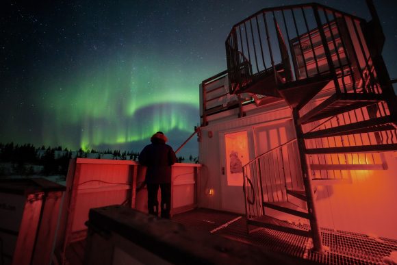 A person stands on a red-lit observation platform beneath a spiral staircase as green aurora borealis glows.