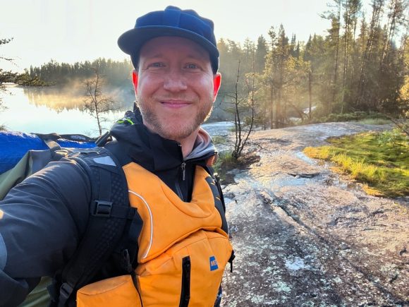 A man wearing an orange life jacket and ball cap takes a selfie on a rocky shoreline in Nopiming Provincial Park.
