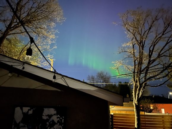 Faint green and purple aurora borealis glows above a house rooftop and bare trees strung with string lights.