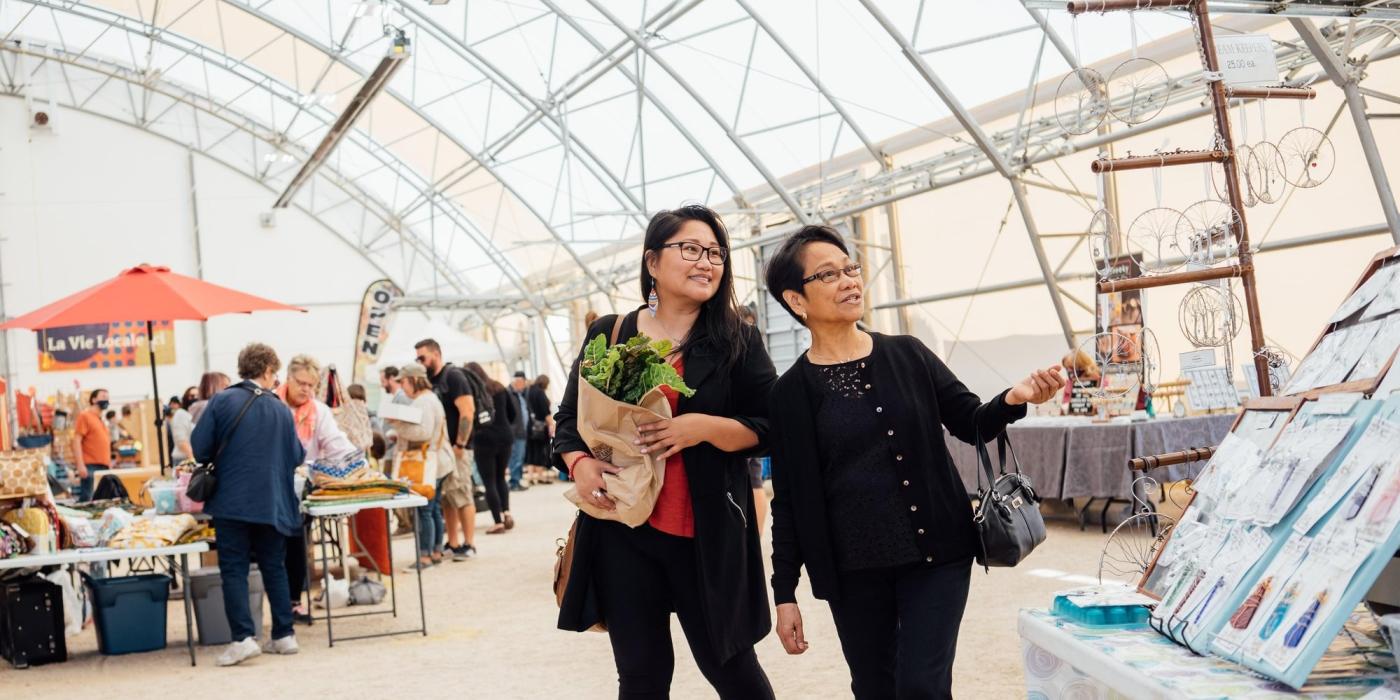 Two people browse St. Norbert Farmer's Market in Winnipeg, one person is carrying a paper bag full of leafy green lettuce.