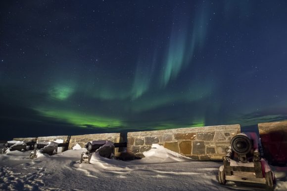 Green aurora borealis arcs across the night sky above a snow-covered stone fort wall lined with old cannons.