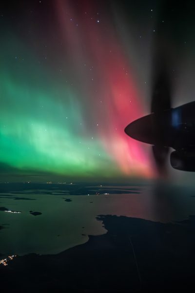 Seen from a plane window past the propeller, red and green aurora borealis glows above a coastline dotted with lights.