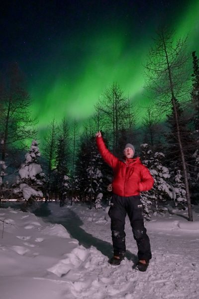 A person in a red jacket raises a celebratory arm while green aurora borealis glowis above snowy spruce trees.