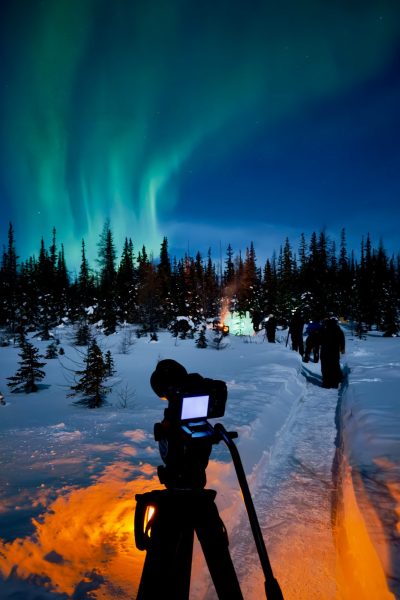 A camera on a tripod films teal-blue aurora borealis over a snowy forest, with an orange campfire glowing in the foreground.