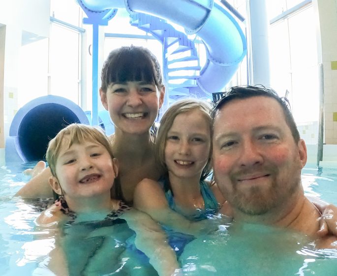 A selfie of a family of four smiles and embraces each other in an indoor pool. A large blue waterslide in the background.