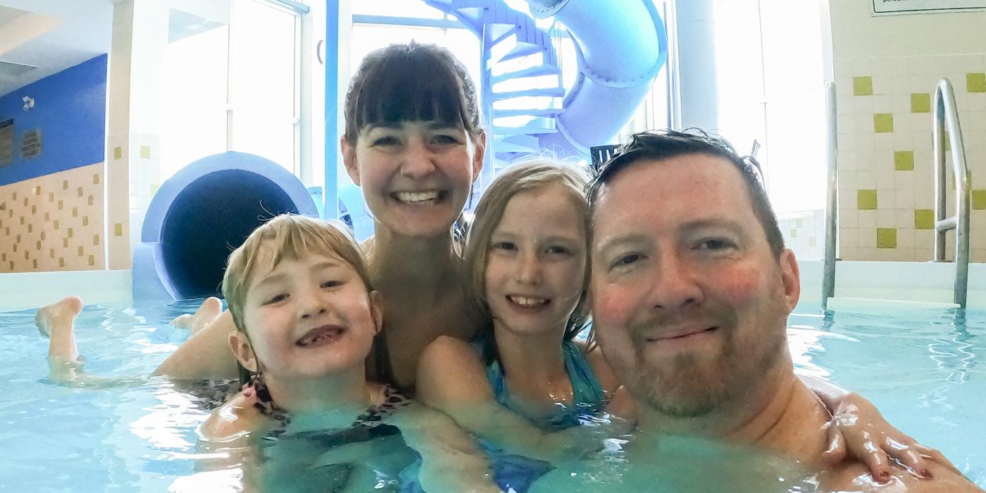 A selfie of a family of four smiles and embraces each other in an indoor pool. A large blue waterslide in the background.
